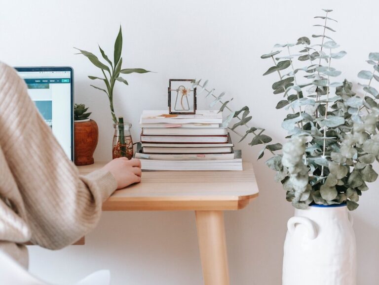 A woman sits at a desk and uses the computer in her cozy gaming room. Plants and a stack of books sit beside her.