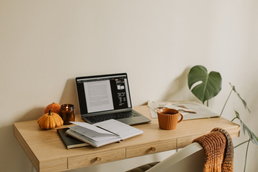 A cozy laptop setup in a gaming room. The desk also has notebooks, coffee, paper, pens, and plants.